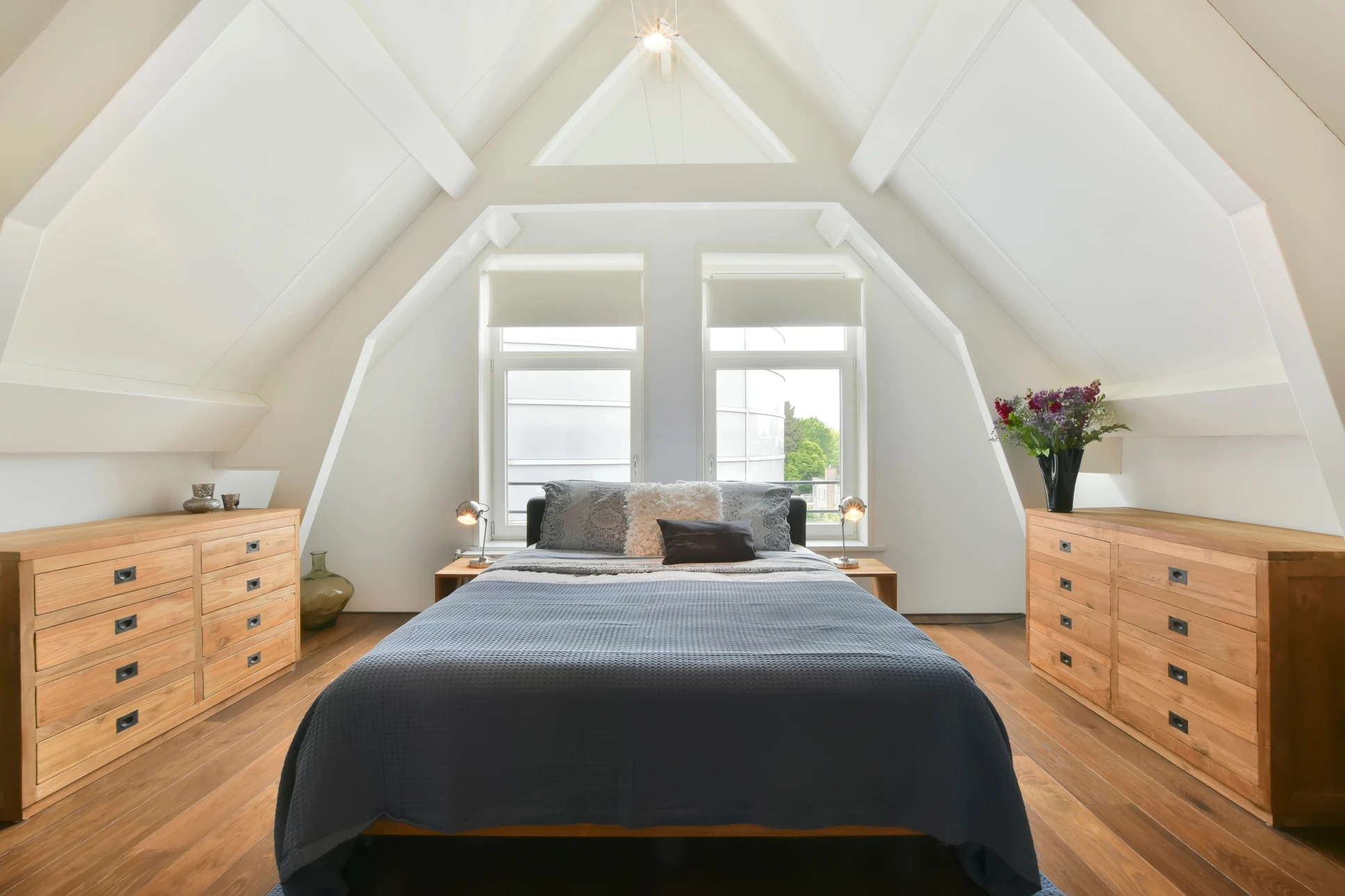 Chambre à coucher sous combles aménagés avec charpente apparente blanche, lit central face à fenêtre double battant
