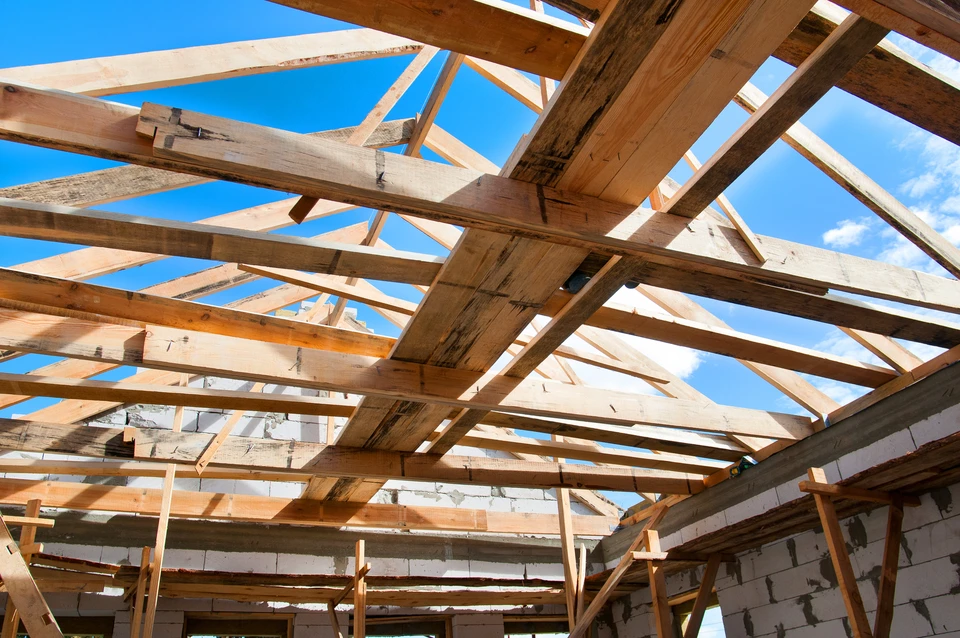 Photographie en plongée d'une charpente traditionnelle en bois massif partiellement assemblée sur un chantier de construction résidentielle, montrant les chevrons, pannes et autres pièces de bois massif taillées avec précision, avec des assemblages artisanaux visibles. Un artisan charpentier peut être présent en bas de l'image pour donner l'échelle, travaillant à la mise en place ou l'ajustement des pièces. L'image doit transmettre la robustesse, la précision et le caractère sur-mesure du travail artisanal de charpenterie traditionnelle.